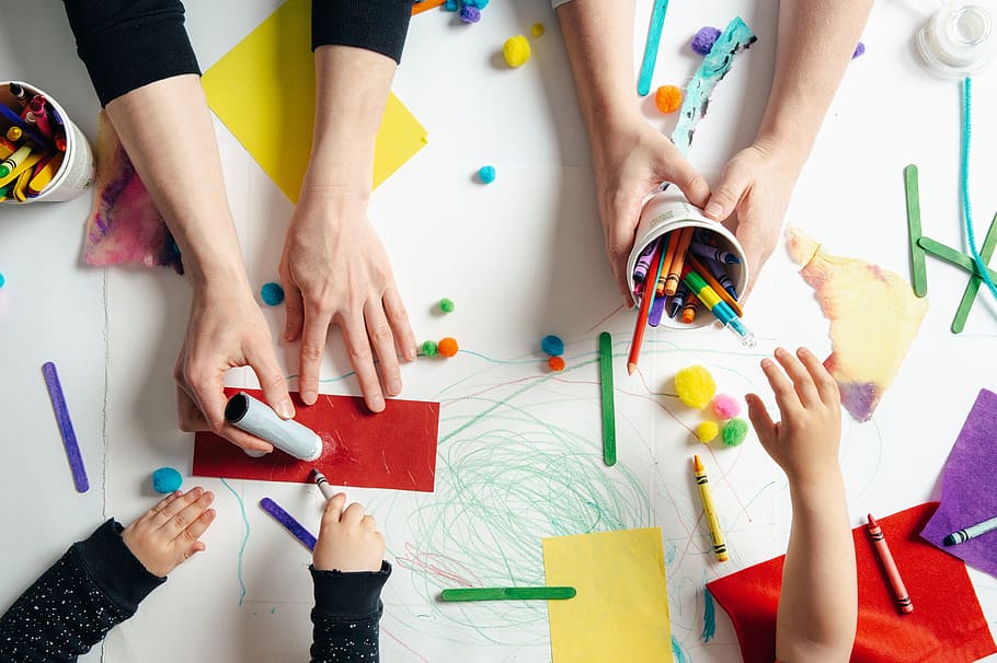 An overhead view of adult and child hands making crafts on a white table covered in crayons, pompoms, lolly sticks and coloured paper.