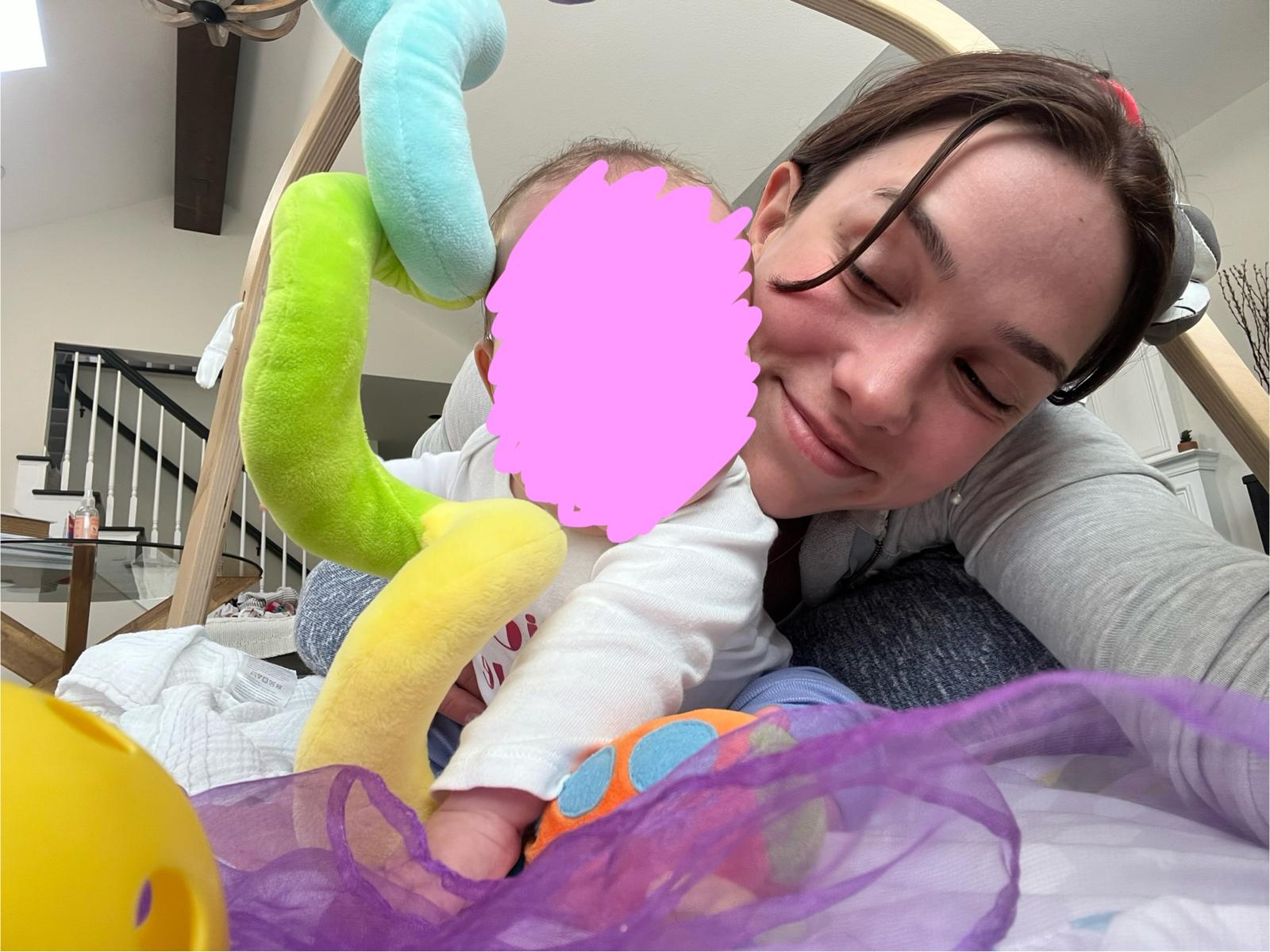 Aurora Carlin Martini lying beside a baby under a wooden play gym, the baby’s face covered by a pink mark drawn over the photograph.