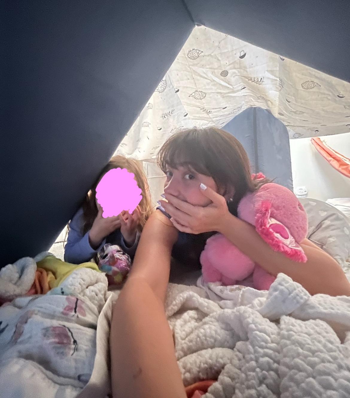 Two children and Aurora Carlin Martini lying inside a blanket fort with soft toys. One child’s face is covered by a pink mark; the other child’s face is uncovered.