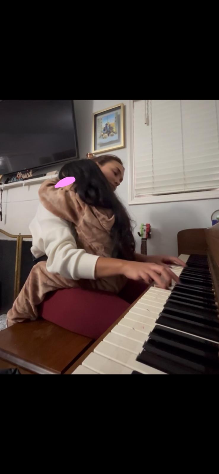 A child in a hooded animal onesie sitting on a piano stool and reaching for the keys, seen from behind, with a small pink mark over the visible part of the face.