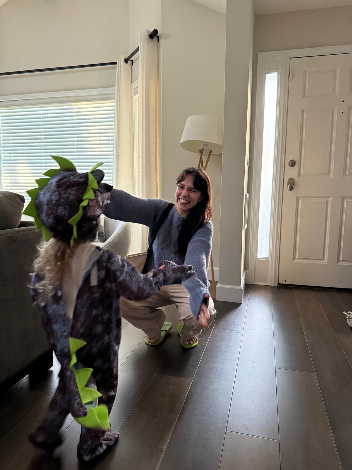 A child in a full dinosaur costume running towards Aurora Carlin Martini, who crouches with both arms open in a living room. The child’s face is inside the costume hood and not visible.