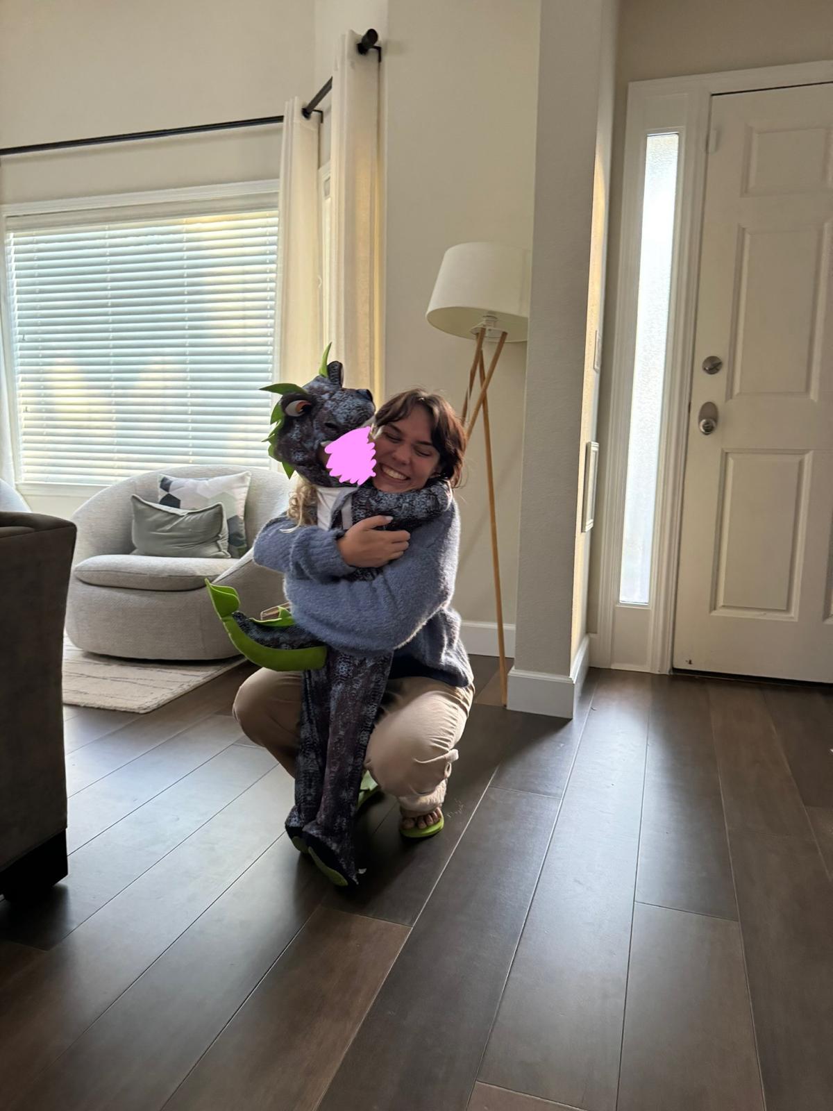 The same child in the dinosaur costume being lifted in a hug by Aurora Carlin Martini, with a small pink mark over the child’s face.