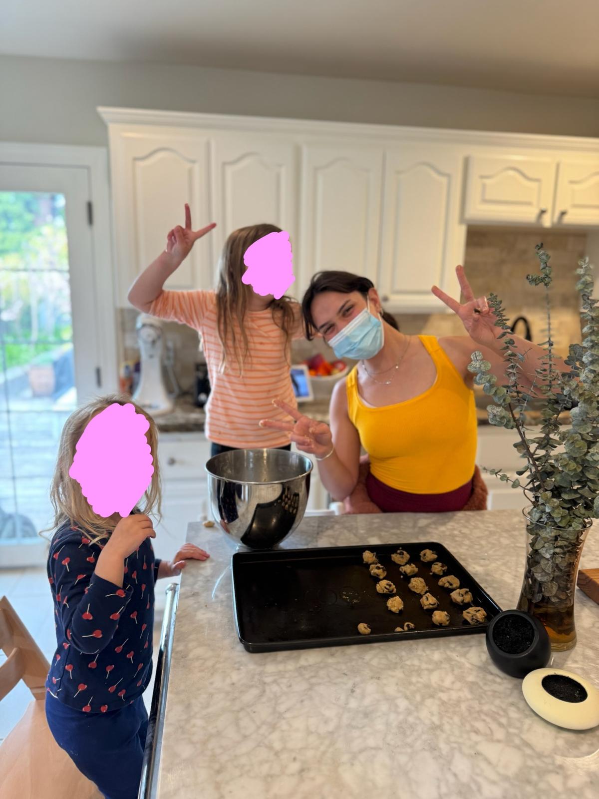 Two children and Aurora Carlin Martini, who is wearing a surgical mask, making peace signs across a kitchen island with a tray of unbaked cookies. Both children’s faces are covered by pink marks.