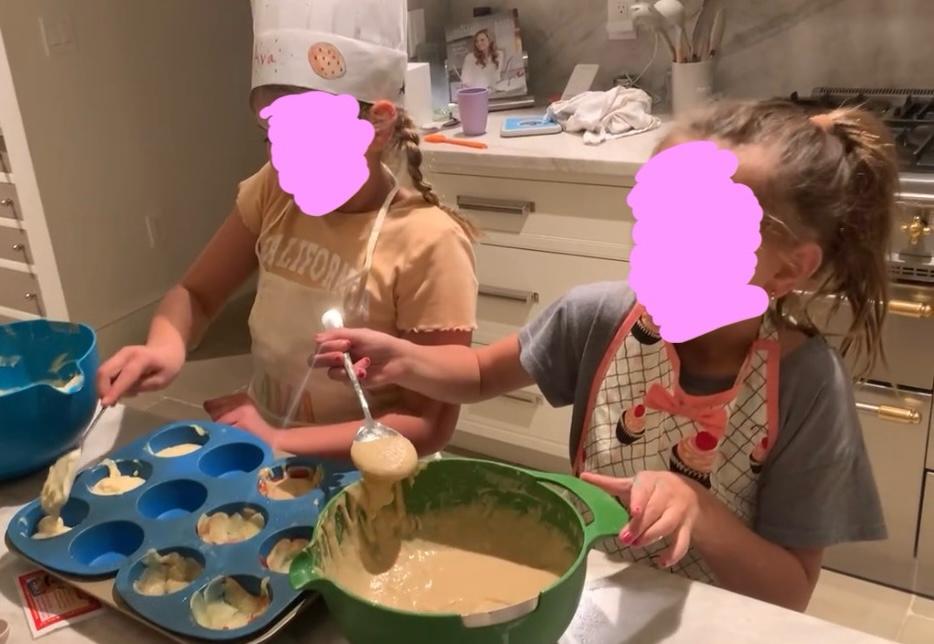 Two children in aprons spooning batter into a muffin tin at a kitchen counter, both faces covered by pink marks. One child wears a white chef’s hat with a name written on it.