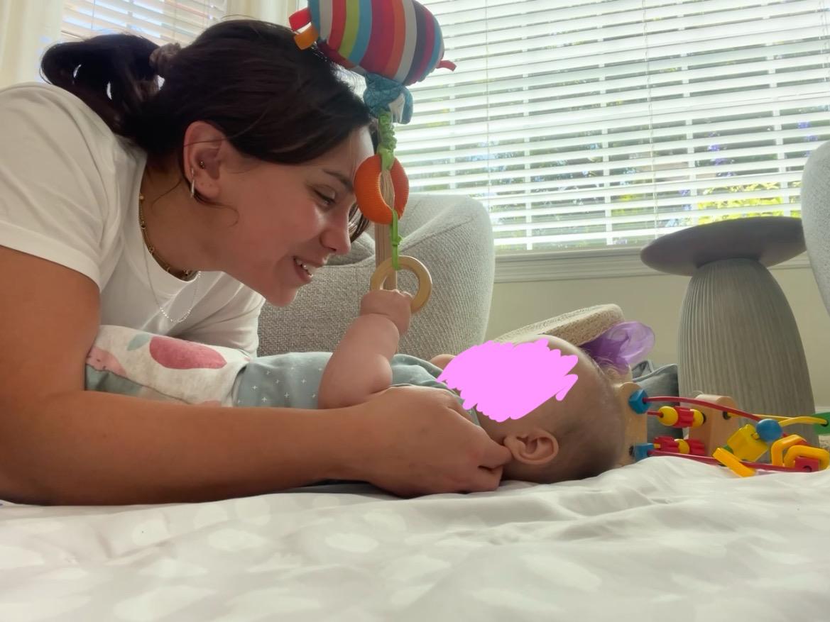 Aurora Carlin Martini lying face to face with a baby on a white bed, holding a wooden toy above them; the baby’s face is covered by a pink mark.