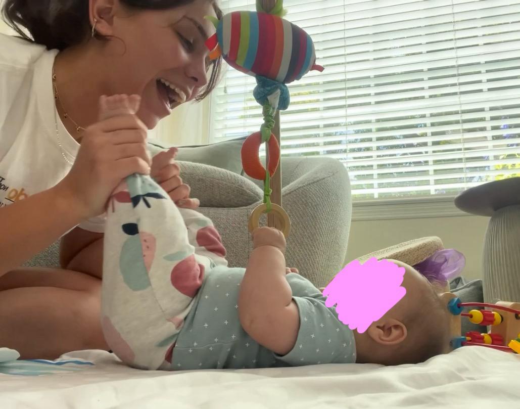 A second frame of the same scene, Aurora Carlin Martini laughing as the baby reaches for the toy, the baby’s face again covered by a pink mark.