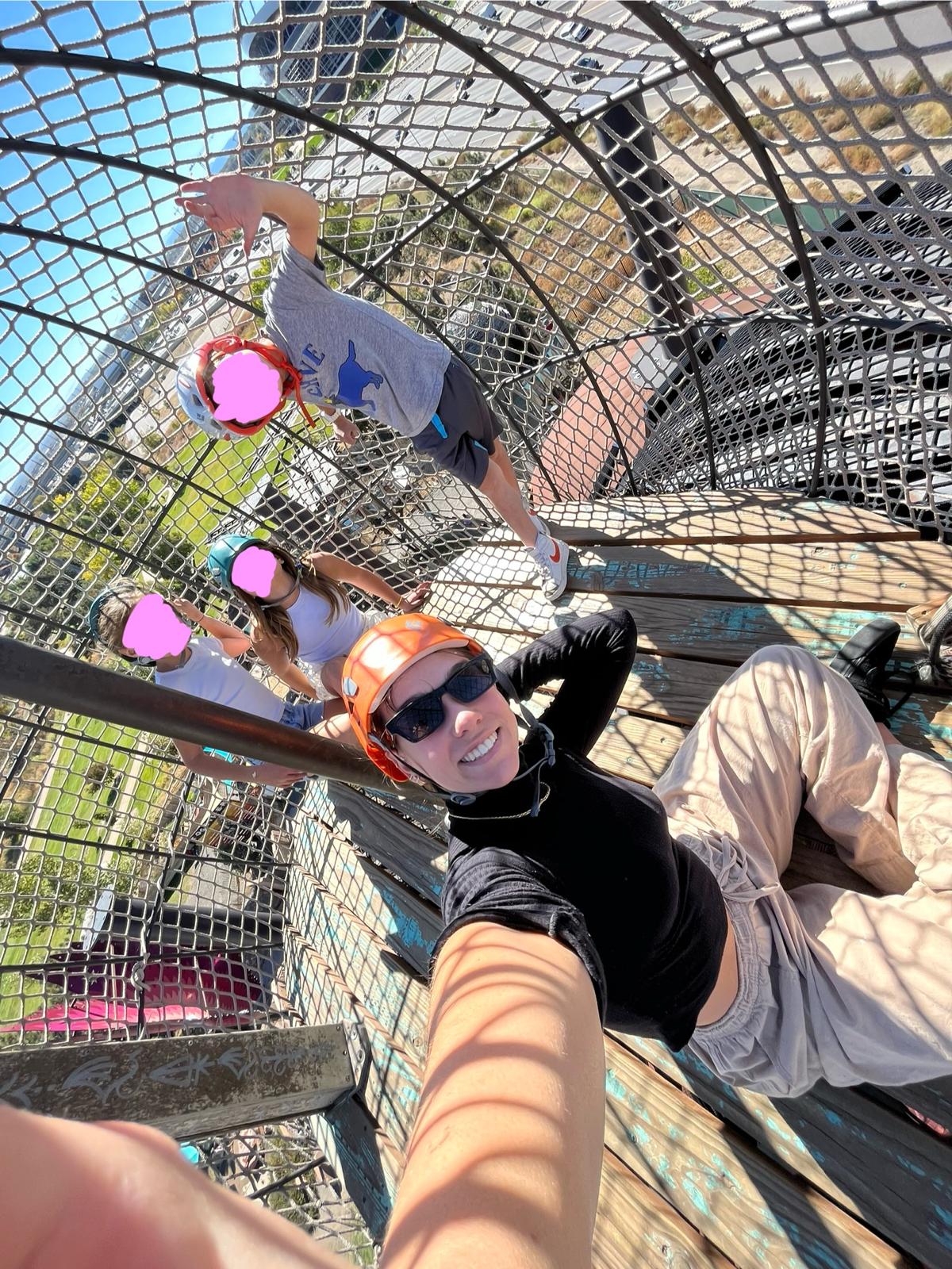 A selfie taken high inside a rope-net climbing structure in a park: Aurora Carlin Martini in a helmet and sunglasses, with three children above and behind her whose faces are covered by pink marks.