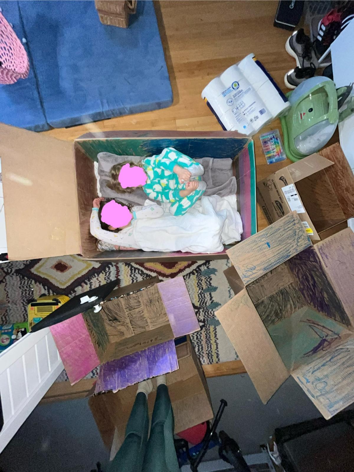 Looking down into a large cardboard box where two children lie side by side under blankets, both faces covered by pink marks, surrounded by flattened painted cardboard on the floor.