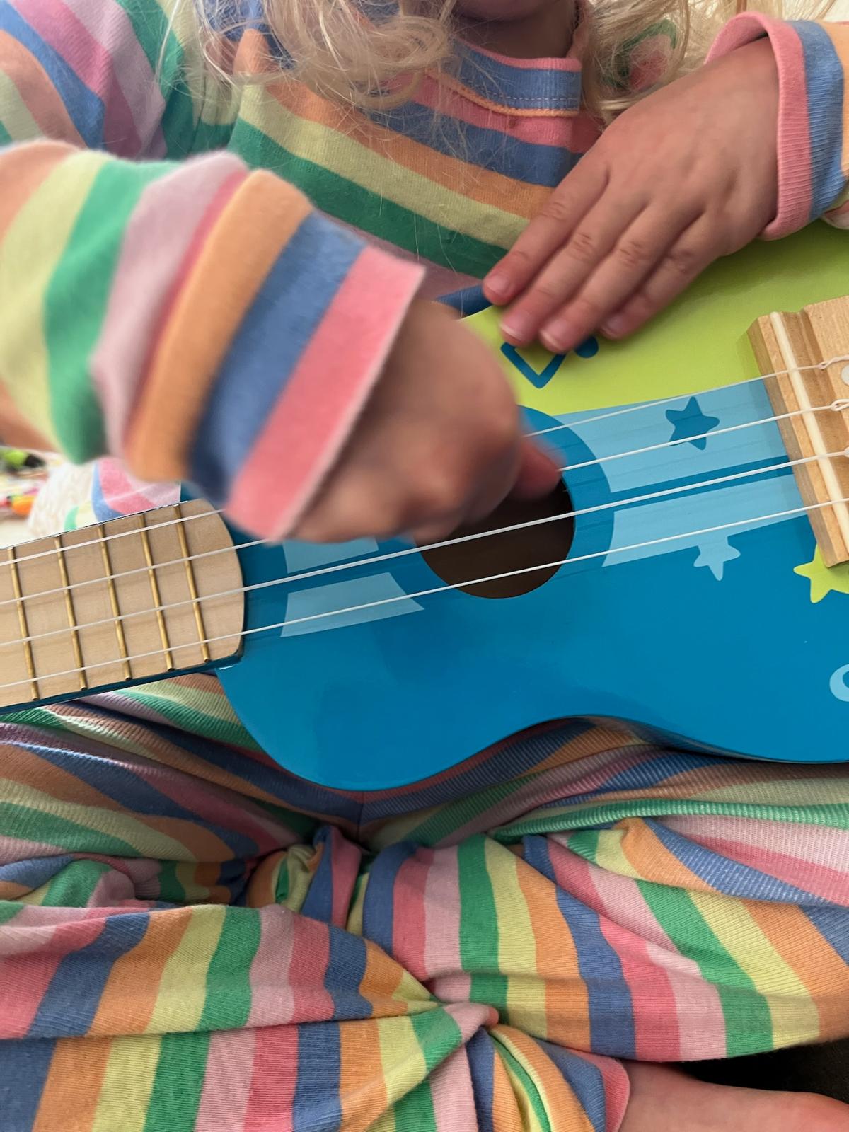 A close view of a small child in rainbow-striped pyjamas strumming a blue toy ukulele, framed from the chin down so no face is visible.