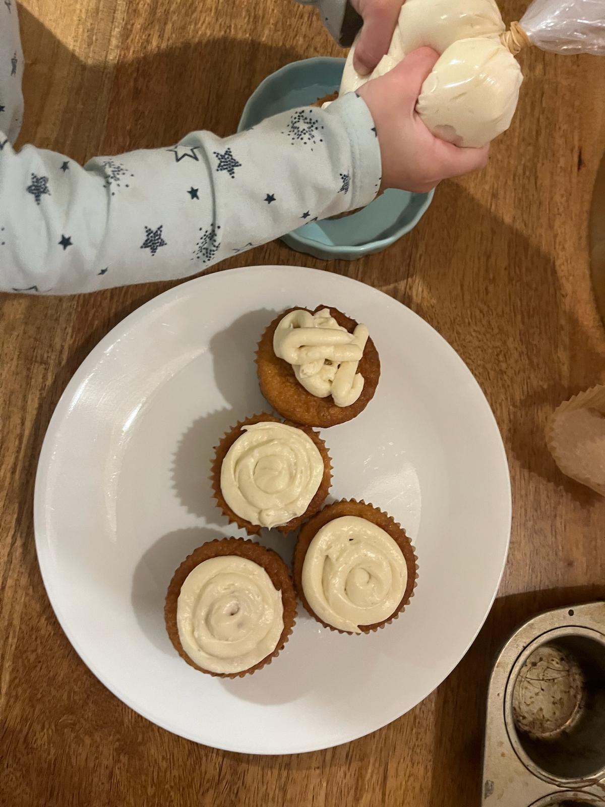 A child’s arm in star-patterned pyjamas squeezing a piping bag of frosting over a plate of four cupcakes on a wooden table.