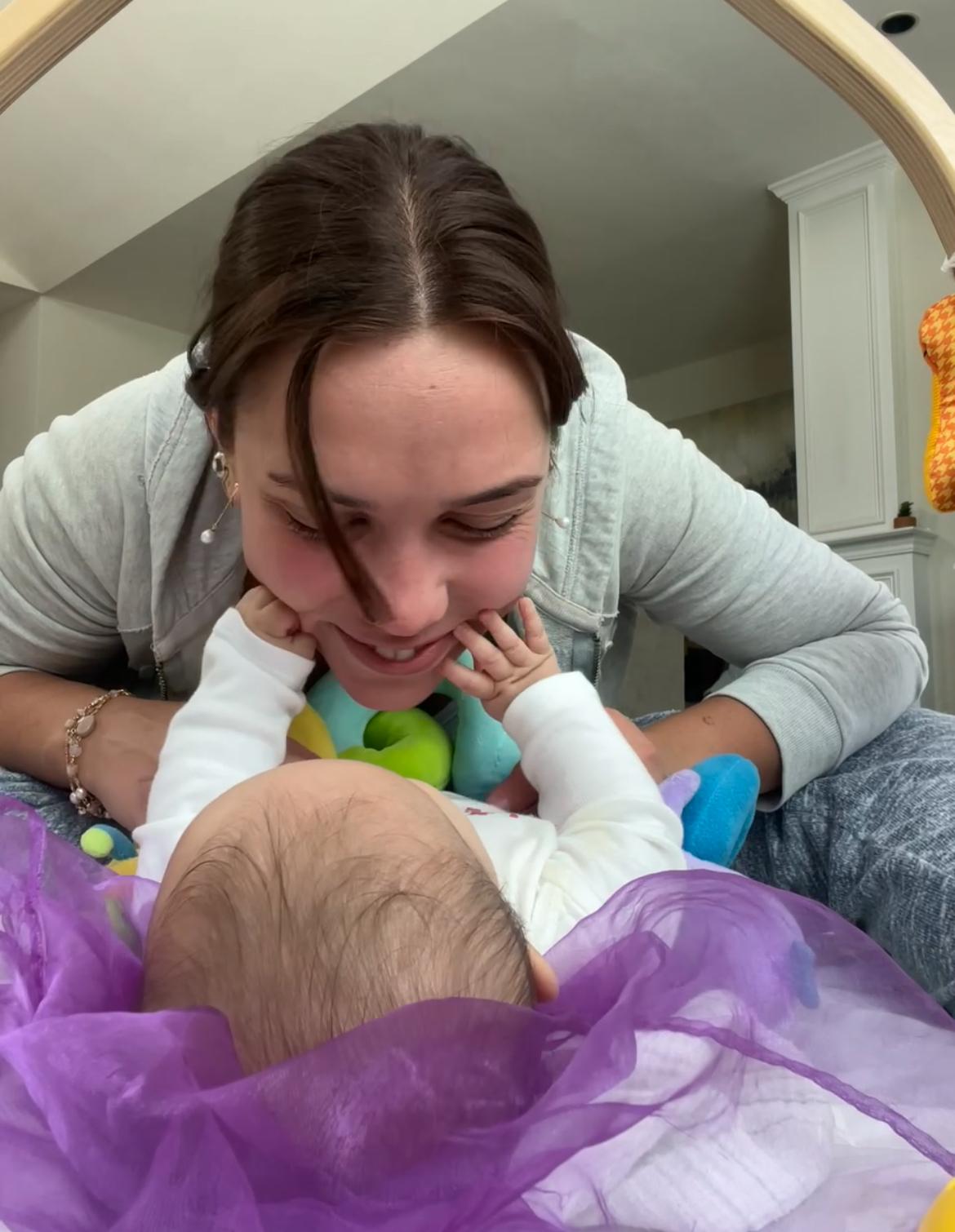 Aurora Carlin Martini leaning over a baby who is lying on a play mat, the baby seen from behind so that only the back of the head is visible.