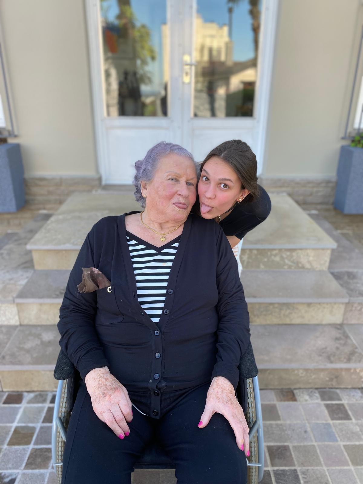 Aurora Carlin Martini leaning over the shoulder of an older woman seated in a wheelchair on a paved terrace, both pulling faces at the camera.