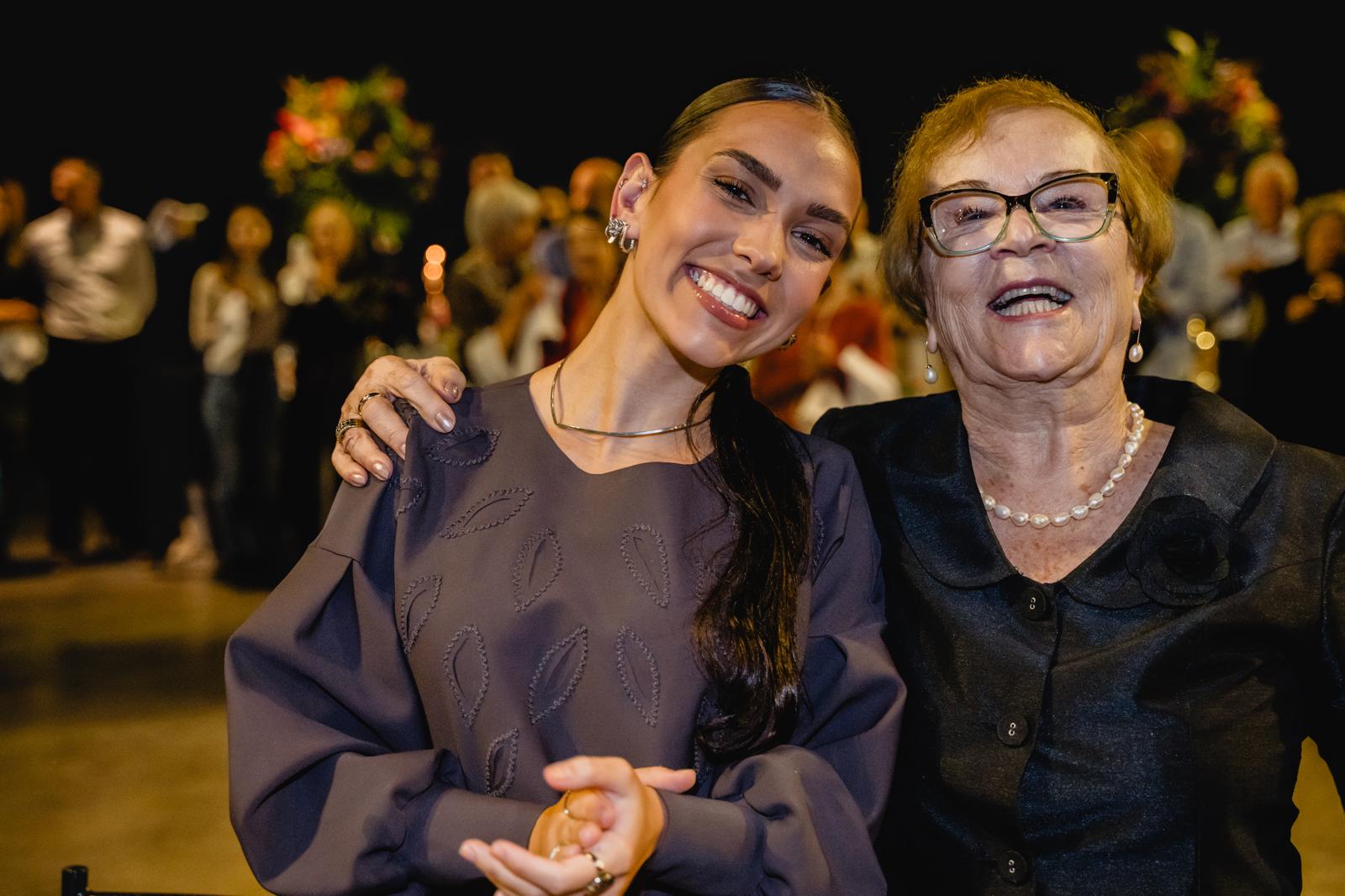 A professionally lit photograph of Aurora Carlin Martini beside a smiling older woman in glasses and pearls, with a blurred crowd and flower arrangements behind them.