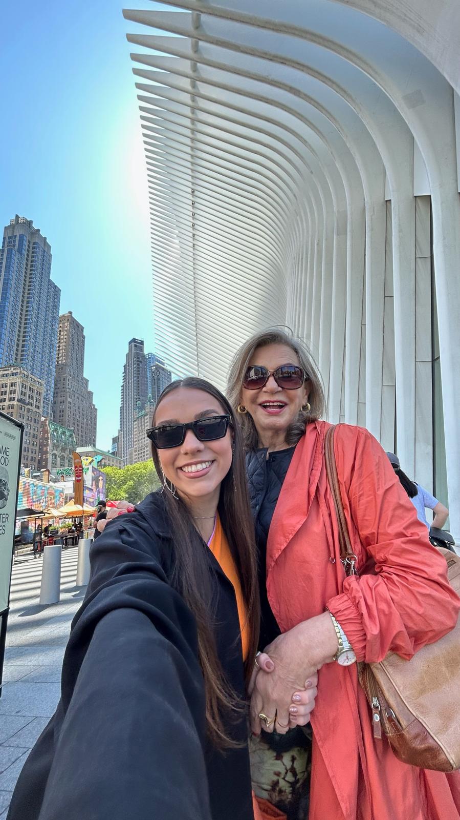 A selfie of Aurora Carlin Martini and an older woman in sunglasses beneath the white ribbed arches of a transit hall, with tall buildings behind them.