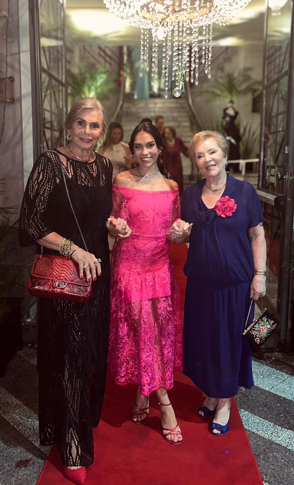 Two older women in evening dress standing either side of a young woman in a pink lace dress on a red carpet, beneath a chandelier.