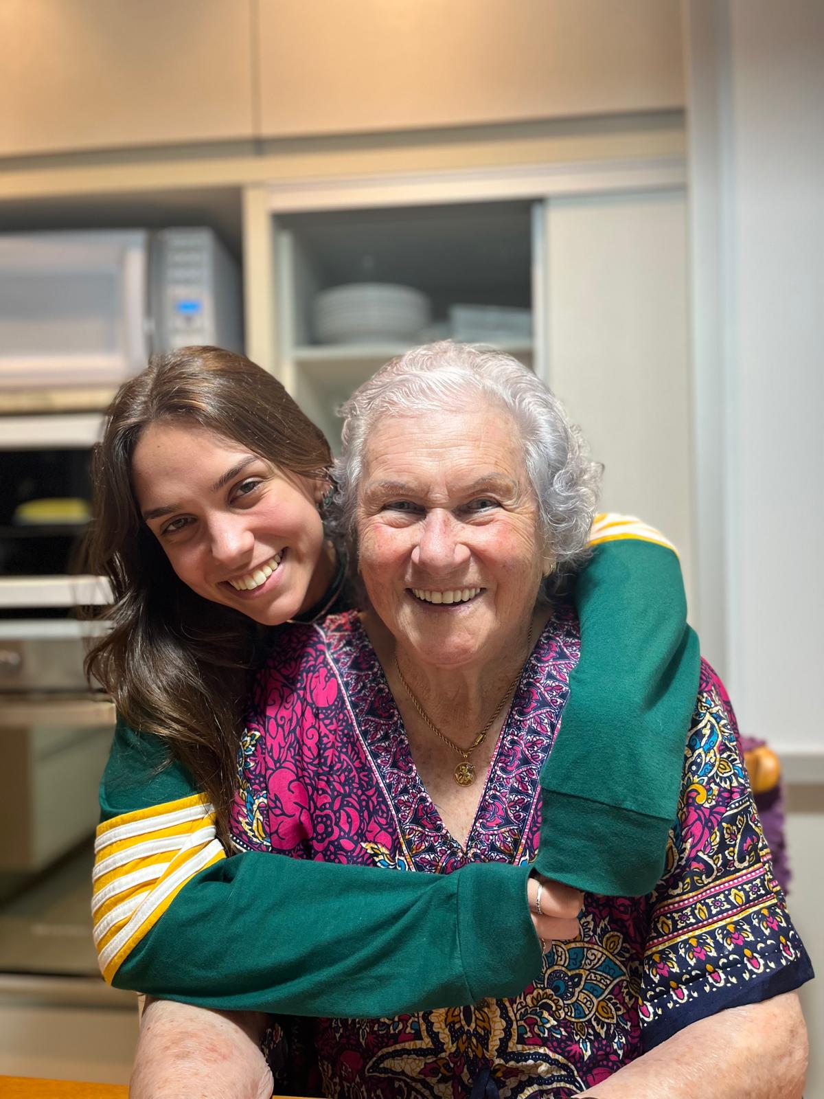 Aurora Carlin Martini with her arms around an older woman in a patterned blouse in a kitchen, both smiling at the camera.