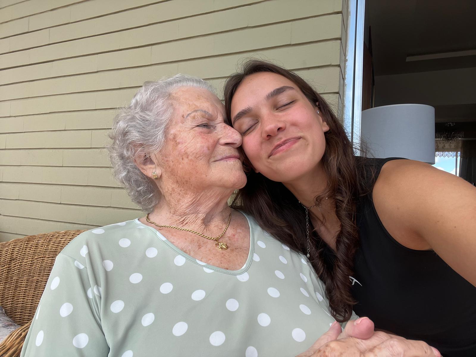Aurora Carlin Martini resting her cheek against the cheek of an older woman in a pale green polka-dot top, seated on a porch beside a wicker chair.