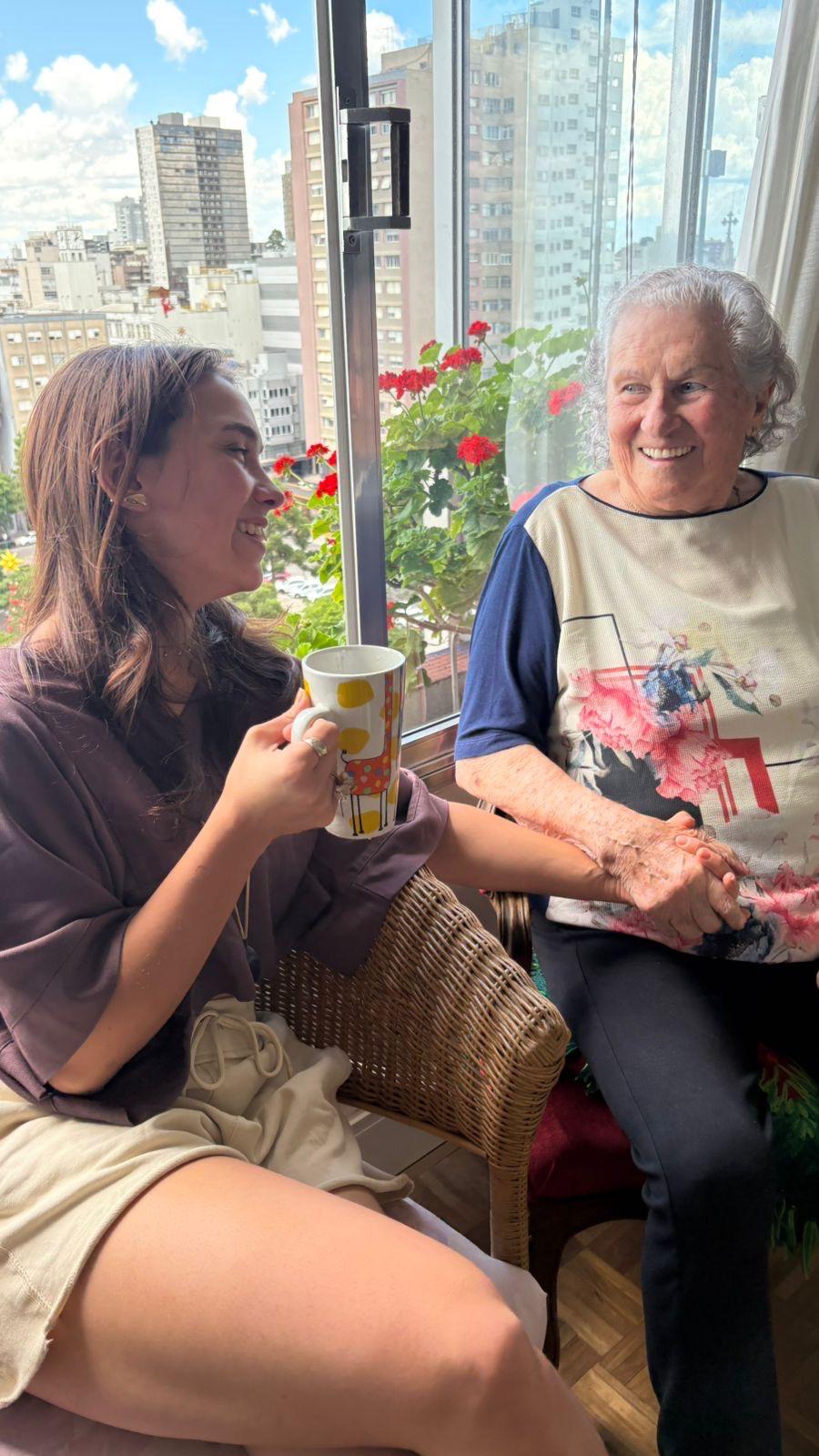 Aurora Carlin Martini holding a mug and laughing beside an older woman in a wicker chair, in front of a window with red geraniums and a city skyline behind.