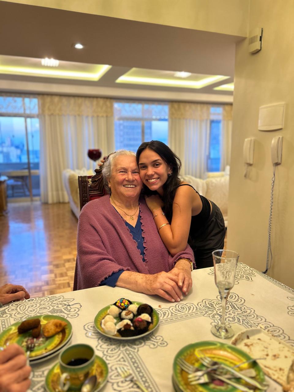 Aurora Carlin Martini leaning down to embrace an older woman seated at a laid dining table with a plate of sweets, in a large room.
