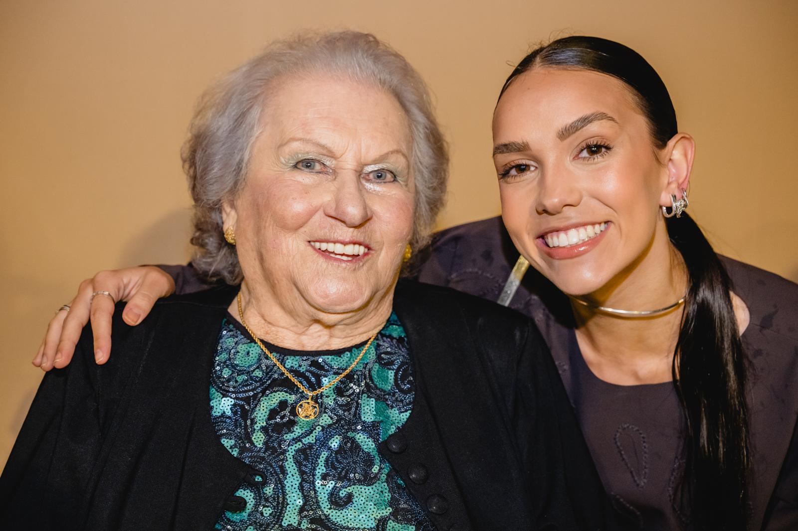 A professionally lit close portrait of an older woman in a sequinned top beside Aurora Carlin Martini, both smiling.