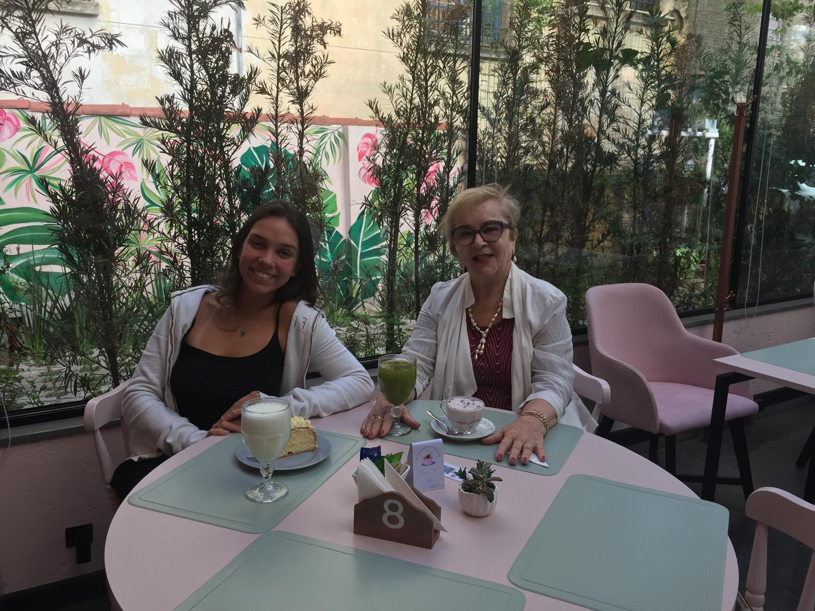 A young woman and an older woman seated at a round café table with drinks and a slice of cake, in front of a painted tropical wall.