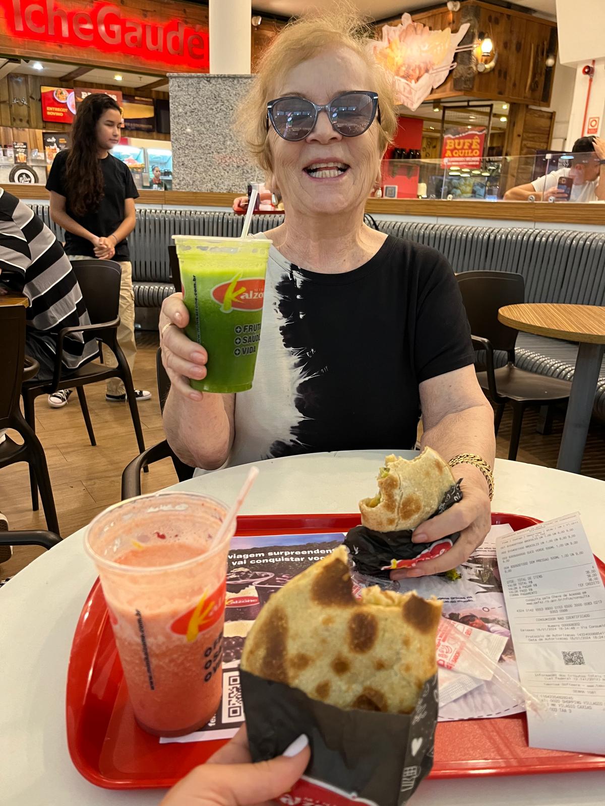 An older woman in sunglasses raising a green juice at a food-court table, with a tray of drinks and pastries in front of her and other diners behind.