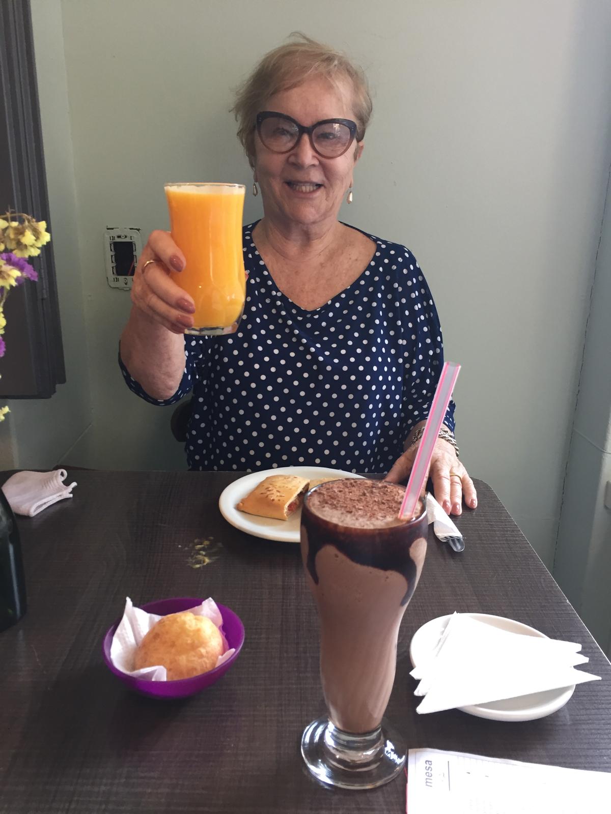 An older woman in glasses and a polka-dot blouse raising a glass of orange juice at a café table set with pastries and a chocolate drink.