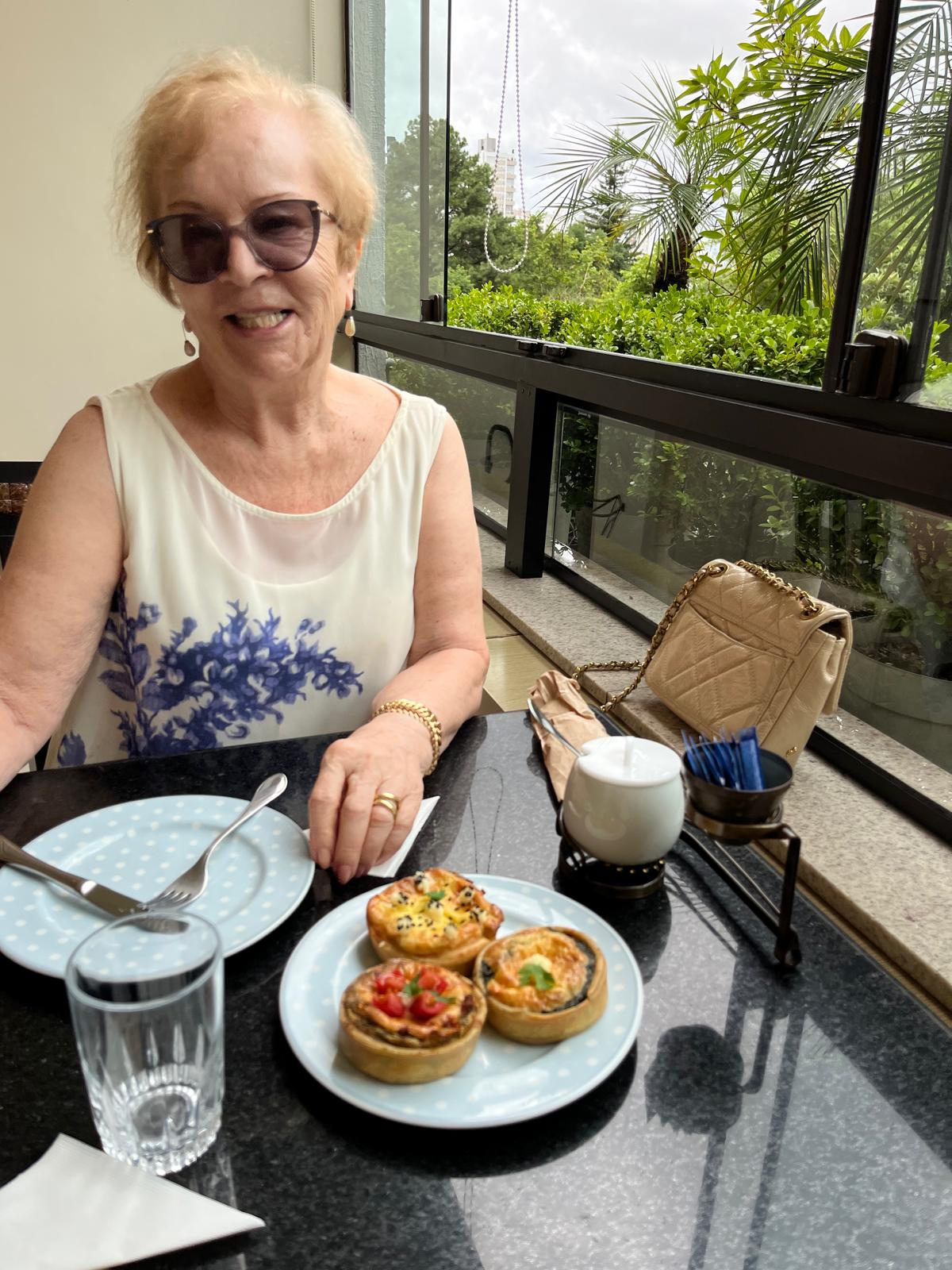 An older woman in sunglasses seated at a dark stone table with a plate of three small quiches, beside a window onto greenery.