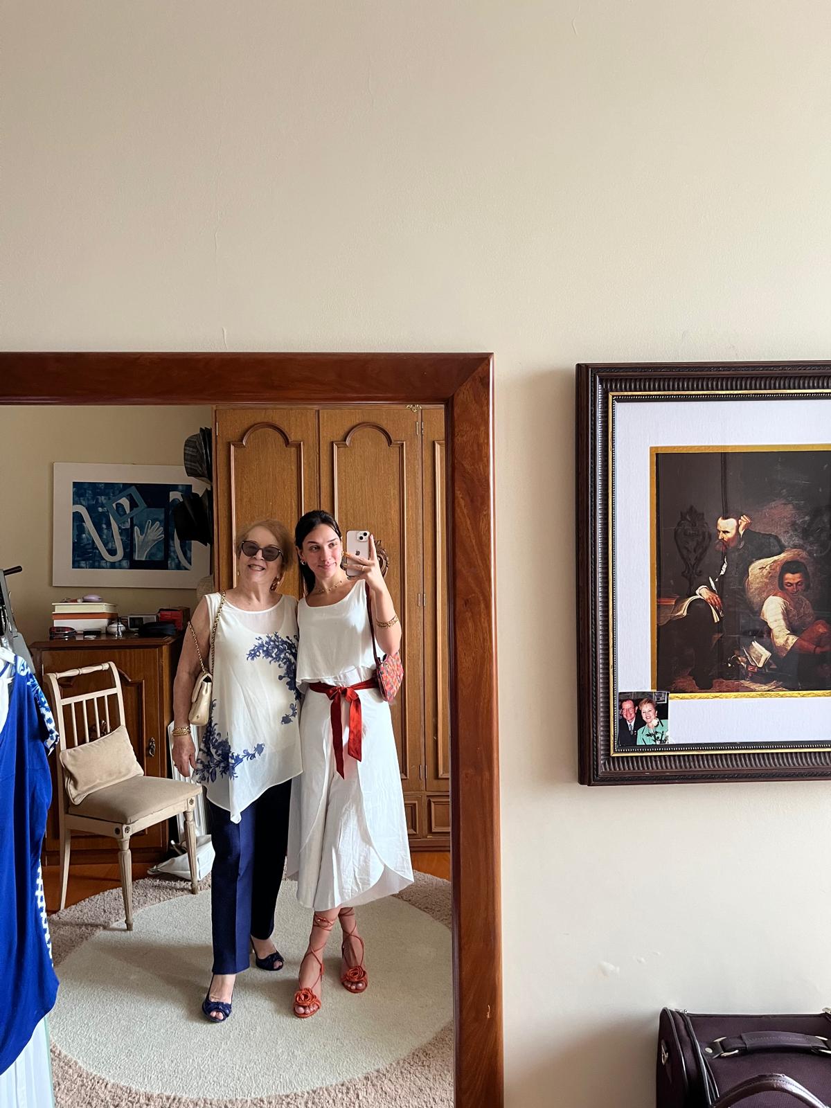 A mirror selfie of Aurora Carlin Martini in a white dress beside an older woman in a blue-and-white blouse, in a bedroom with a framed painting on the wall.