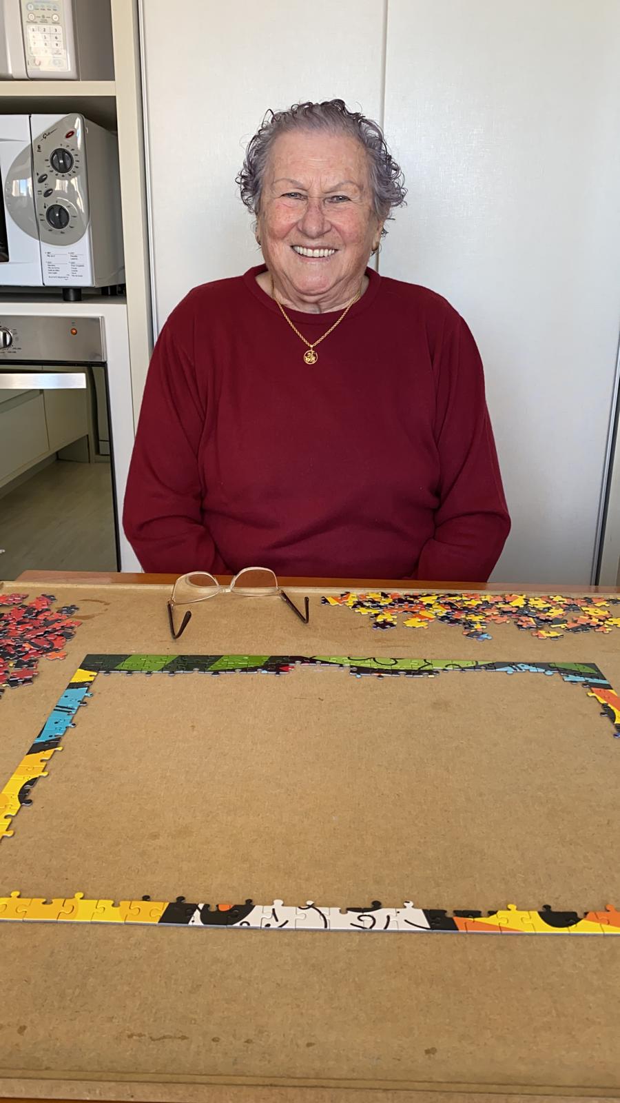 An older woman in a red jumper smiling behind a board holding a partly assembled jigsaw puzzle, her glasses resting on the board.