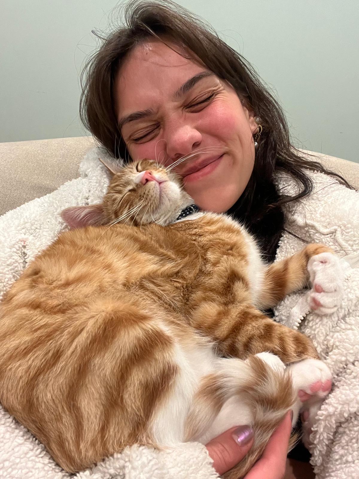 Aurora Carlin Martini indoors holding a ginger-and-white cat against her cheek, both with their eyes closed.