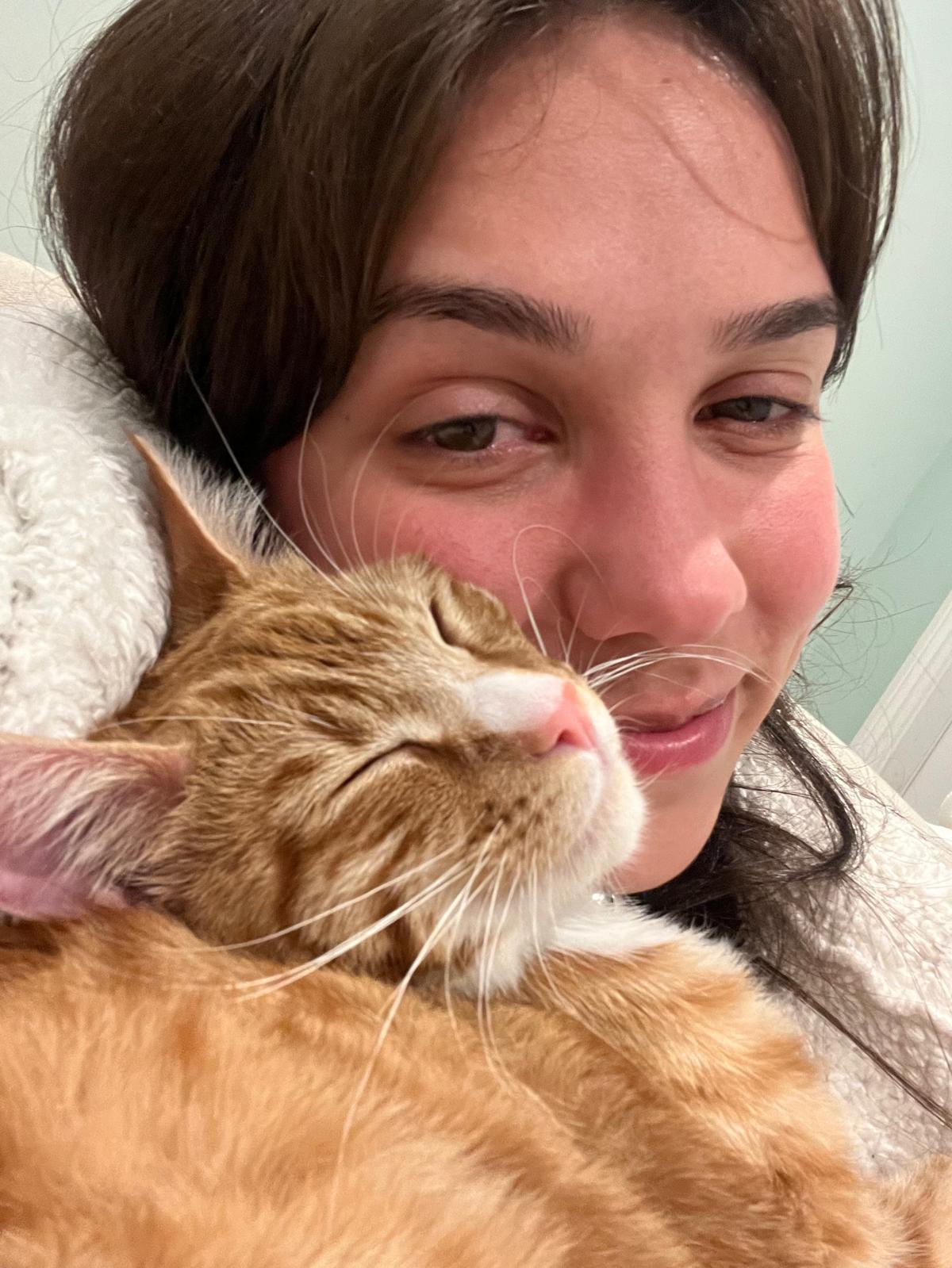 Aurora Carlin Martini in close-up with the same ginger-and-white cat held against her shoulder, against a pale green wall.