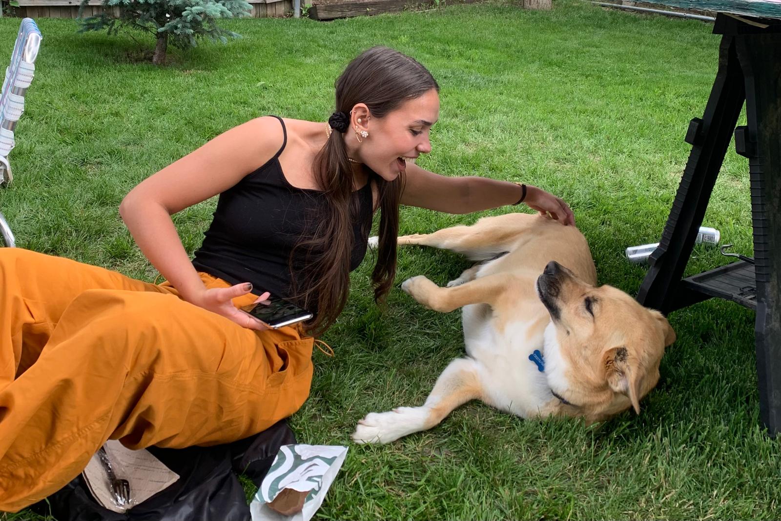 A young woman kneeling on a lawn, one hand on the chest of a tan dog lying on its back beside her; a second person in an orange jacket is partly in frame.