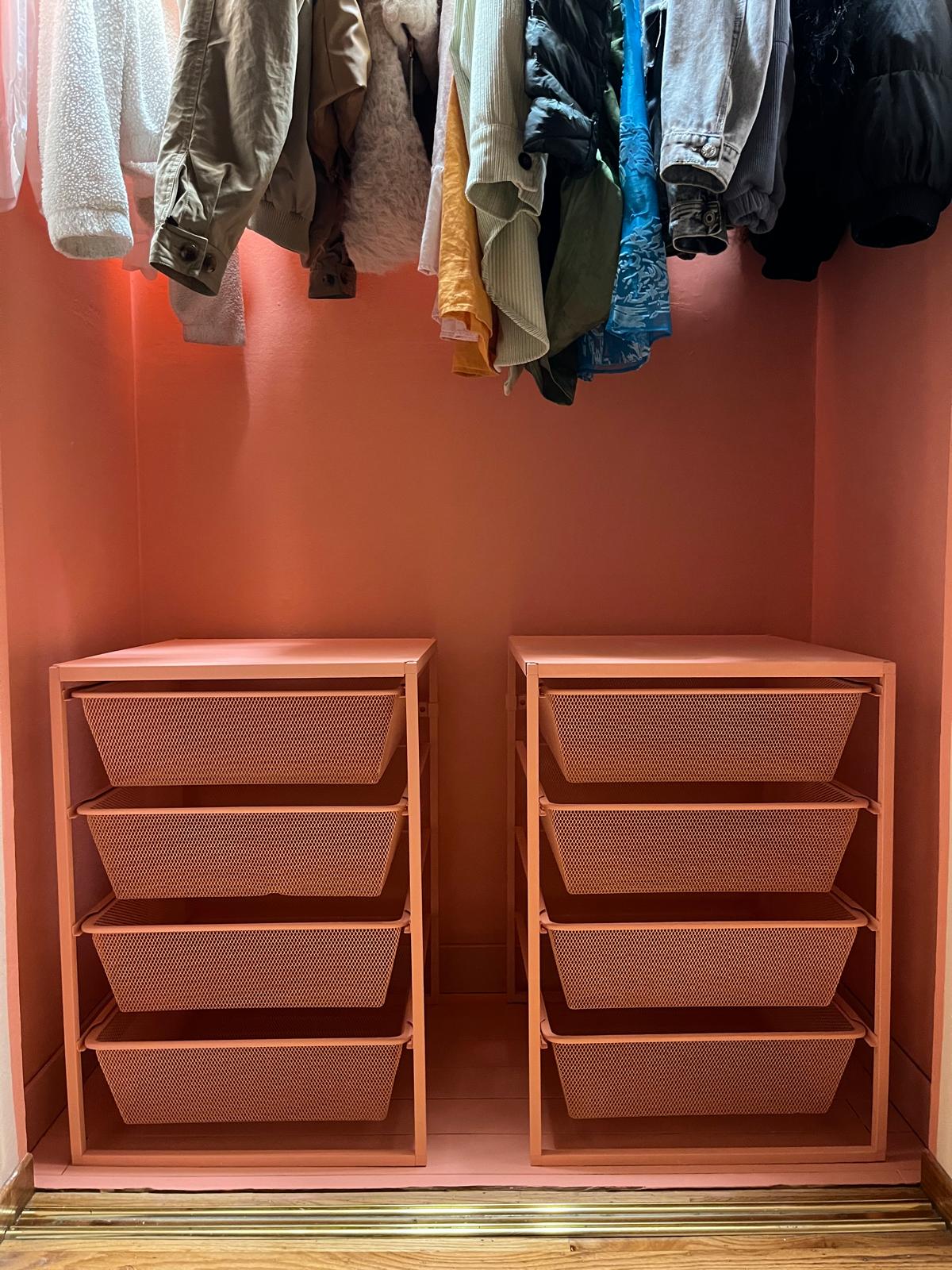 A closet niche painted terracotta, holding two salmon-coloured units of mesh drawers, with coats hanging on a rail above them.