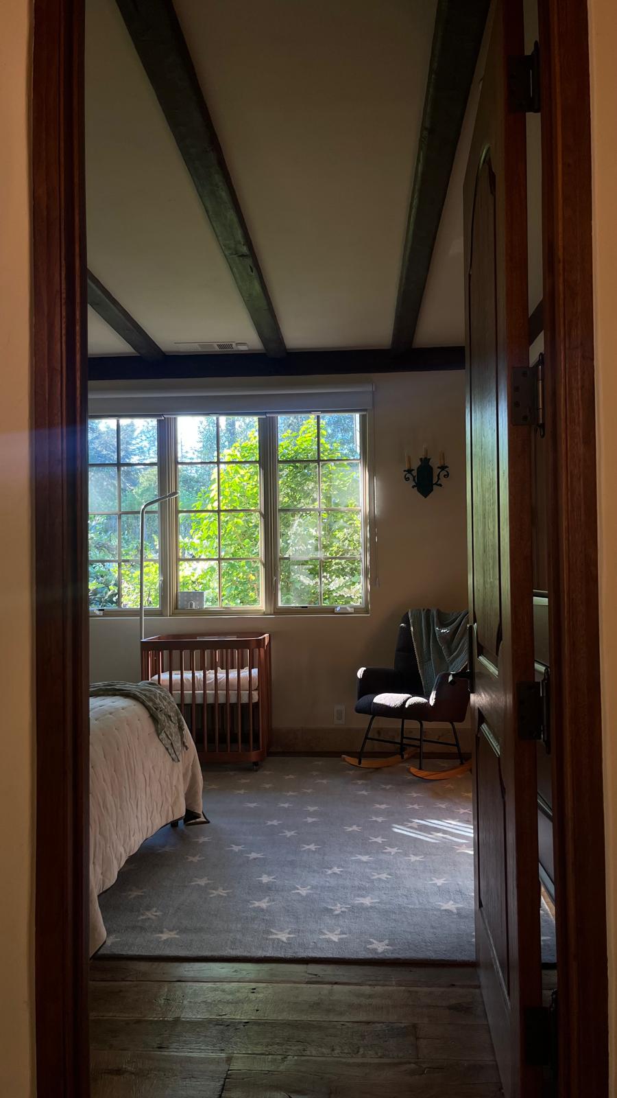 A nursery seen through an open panelled door: a wooden cot under a window, a rocking chair with a blanket over it, a star-patterned rug and a beamed ceiling.