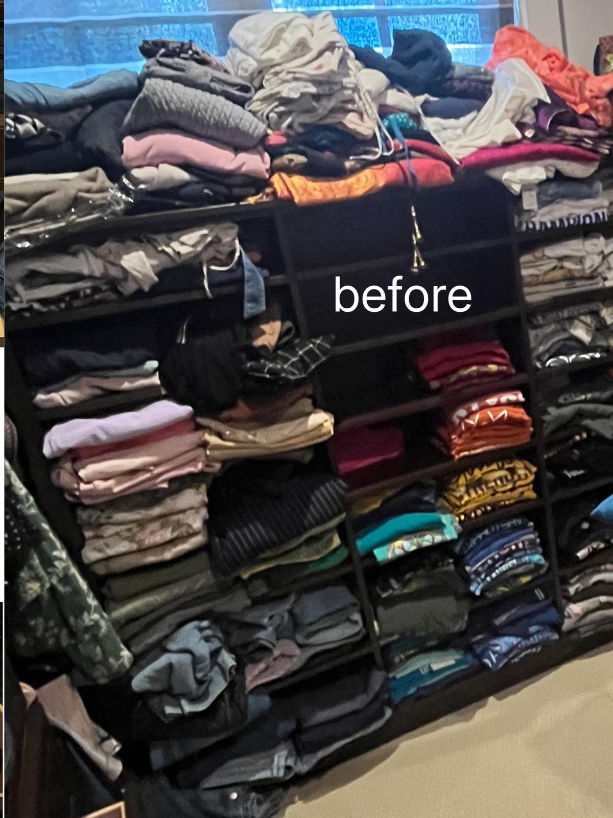 A dark cube shelving unit overflowing with clothes piled loose on every shelf and heaped across the top.