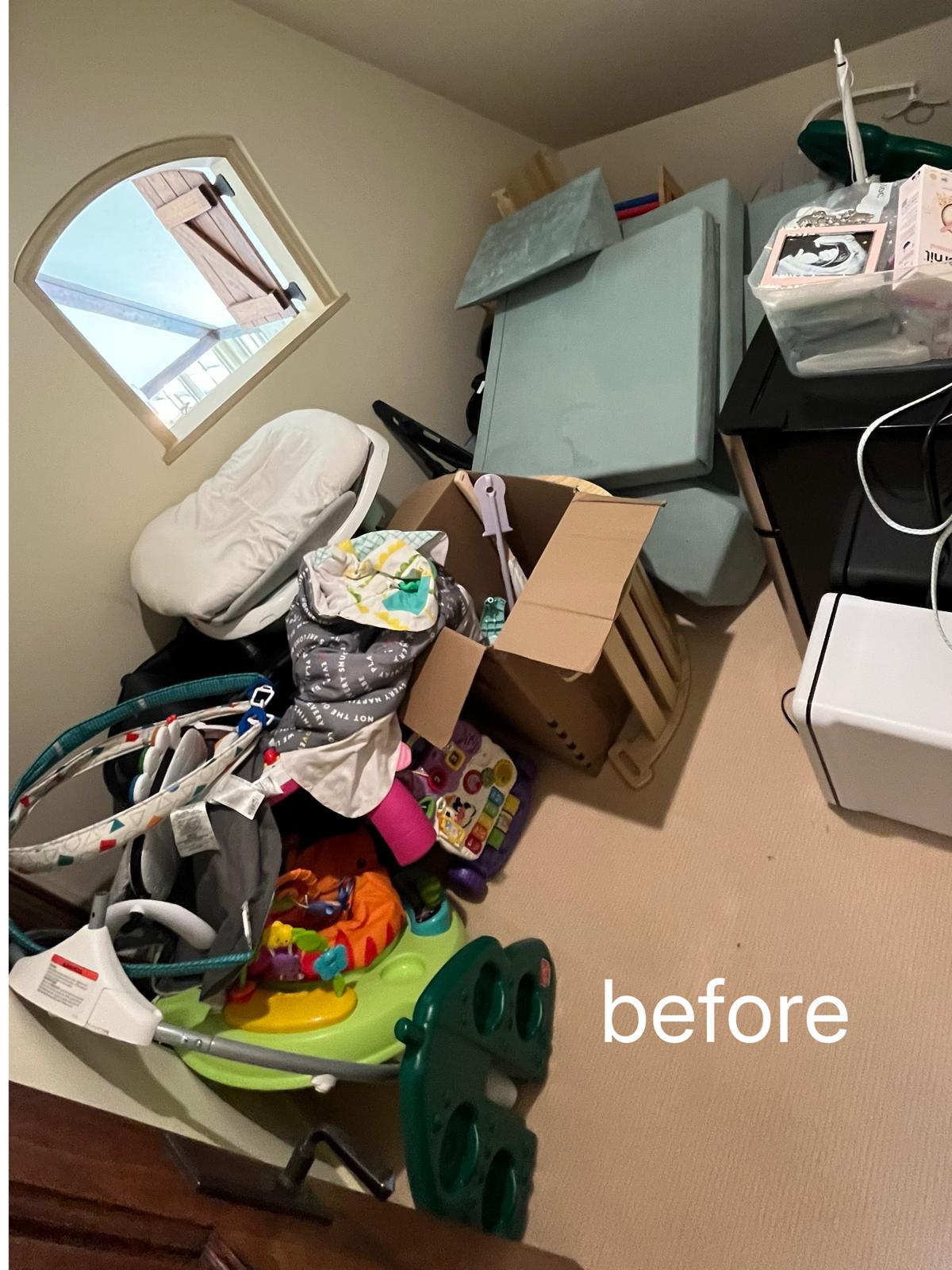 A small storage room with baby equipment, folded play mats, cardboard boxes and a bouncer piled against the walls and across the floor.