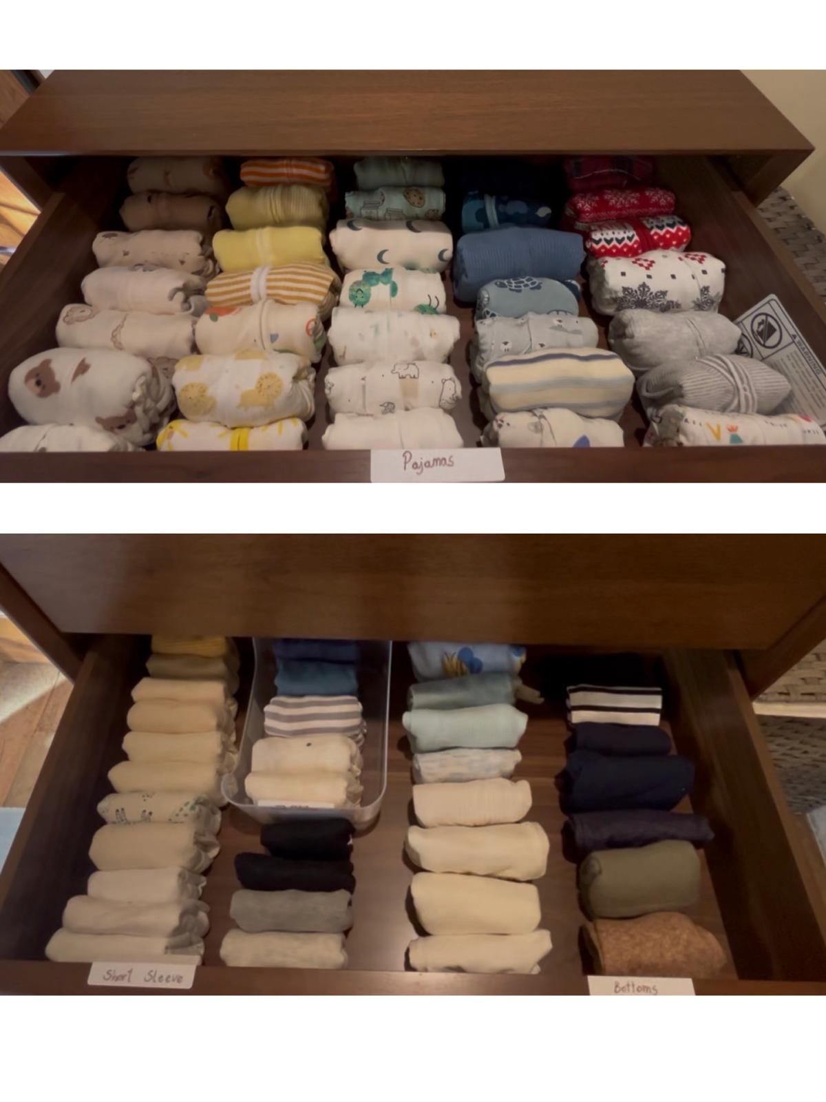 Two open wooden drawers of baby clothes, each garment rolled and filed upright, with handwritten labels reading “Pajamas”, “Short Sleeve” and “Bottoms”.