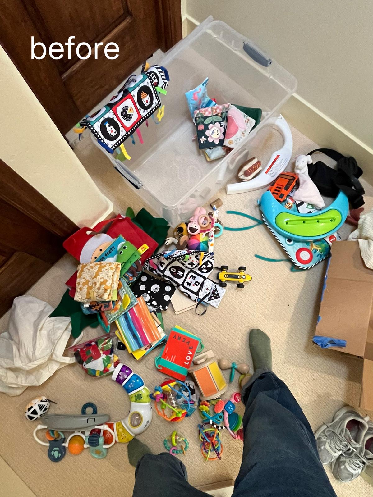Baby toys, books and rattles spread across a hallway carpet beside an emptied storage box, with a person’s legs and feet at the bottom of the frame.