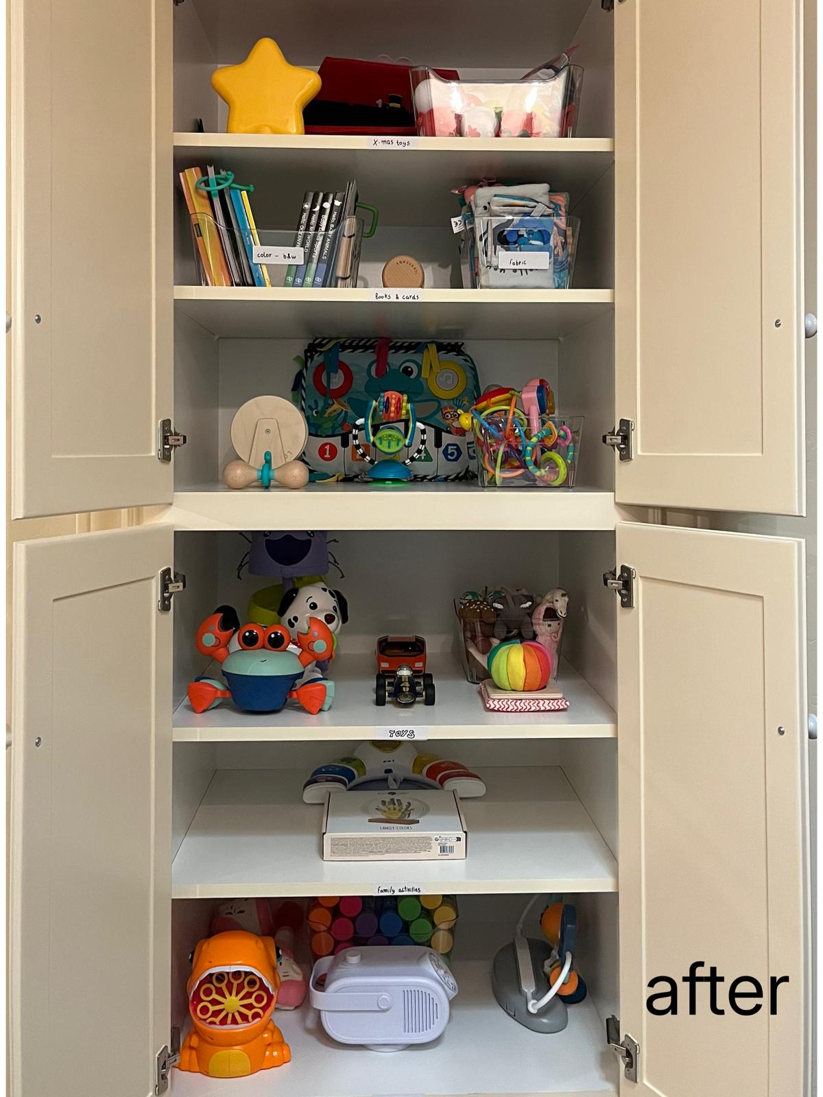 A tall cream cabinet with both doors open, its six shelves holding sorted toys, books and activity boxes, each shelf edge carrying a printed label.